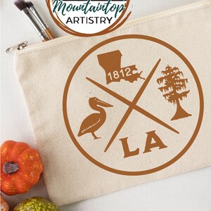 May include: A brown canvas zippered pouch with a brown circle design featuring a pelican, a tree, the state of Louisiana, and the text "1812" and "LA". The pouch is on a white surface with two small orange pumpkins and one green pumpkin.