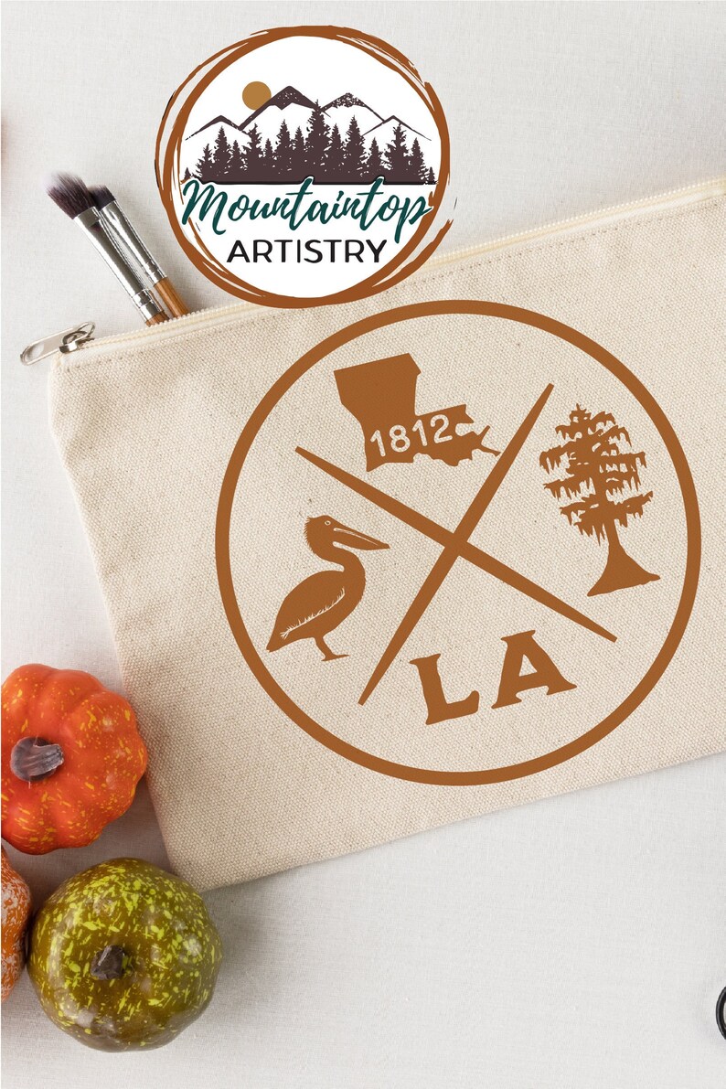 May include: A brown canvas zippered pouch with a brown circle design featuring a pelican, a tree, the state of Louisiana, and the text "1812" and "LA". The pouch is on a white surface with two small orange pumpkins and one green pumpkin.