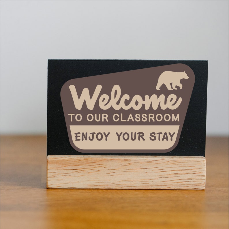 Puede incluir: Un letrero negro de pizarra con una base de madera, que presenta un letrero de bienvenida marr&oacute;n y beige con una silueta de oso. El letrero dice "Bienvenido a nuestra aula, Disfruta de tu estancia."
