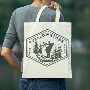 May include: A white tote bag with a gray graphic of an elk and trees. The graphic is in the shape of a hexagon with the text "Yellowstone National Park" around the outside.