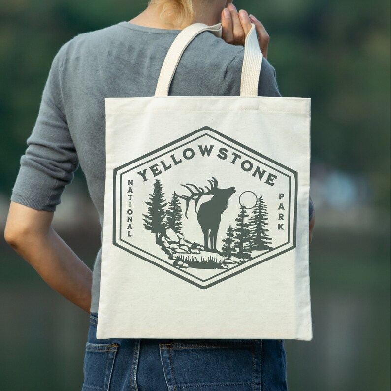 May include: A white tote bag with a gray graphic of an elk and trees. The graphic is in the shape of a hexagon with the text "Yellowstone National Park" around the outside.