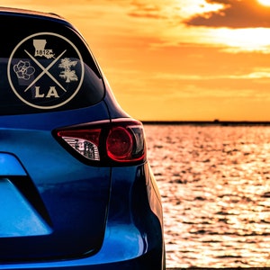 May include: A blue car with a gold sticker on the back window. The sticker features a magnolia flower, a cypress tree, and the text "LA 1812". The car is parked near a body of water with a sunset in the background.