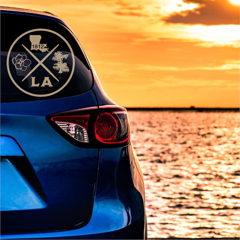 May include: A blue car with a gold sticker on the back window. The sticker features a magnolia flower, a cypress tree, and the text "LA 1812". The car is parked near a body of water with a sunset in the background.