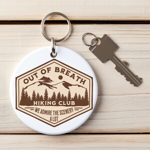 May include: White round keychain with a brown hexagonal logo that says "Out of Breath Hiking Club - We Admire the Scenery a Lot". The logo features a mountain range with a sun and trees.