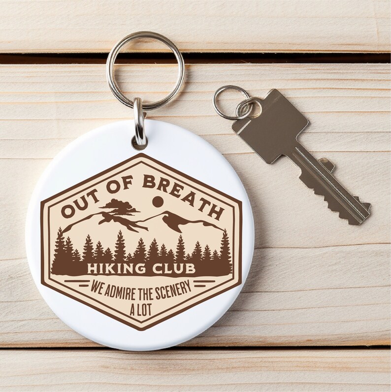 May include: White round keychain with a brown hexagonal logo that says "Out of Breath Hiking Club - We Admire the Scenery a Lot". The logo features a mountain range with a sun and trees.