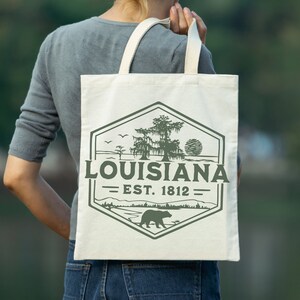 May include: A white canvas tote bag with a green and brown graphic design. The graphic features a bear, a tree, and the text "Louisiana - Est. 1812".