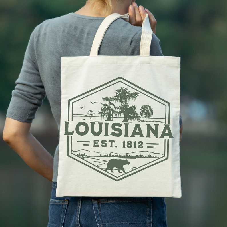 May include: A white canvas tote bag with a green and brown graphic design. The graphic features a bear, a tree, and the text "Louisiana - Est. 1812".