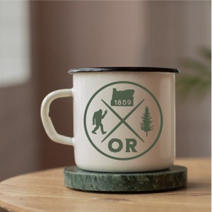 May include: White enamel mug with a black rim and a green graphic of a stylised tree, a Bigfoot, and the state of Oregon with the text "OR" and "1859".
