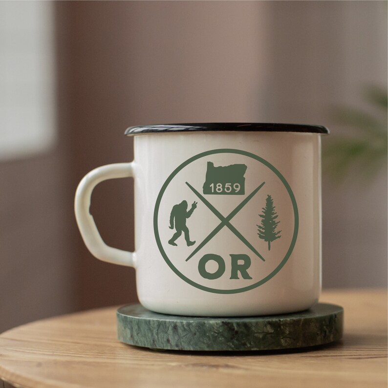 May include: White enamel mug with a black rim and a green graphic of a stylised tree, a Bigfoot, and the state of Oregon with the text "OR" and "1859".