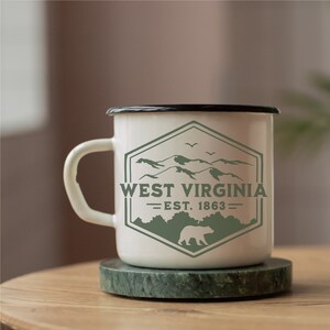 May include: White enamel mug with a black rim and handle. The mug features a green logo with a mountain range, a bear, and the text "West Virginia - Est. 1863 - ".