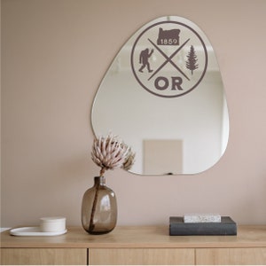 May include: A brown, irregularly shaped mirror with a sticker on it. The sticker features a stylised graphic of a tree, a Bigfoot, and the state of Oregon. The text "OR" and "1859" are also included in the sticker. A vase with a dried flower arrangement is in front of the mirror.