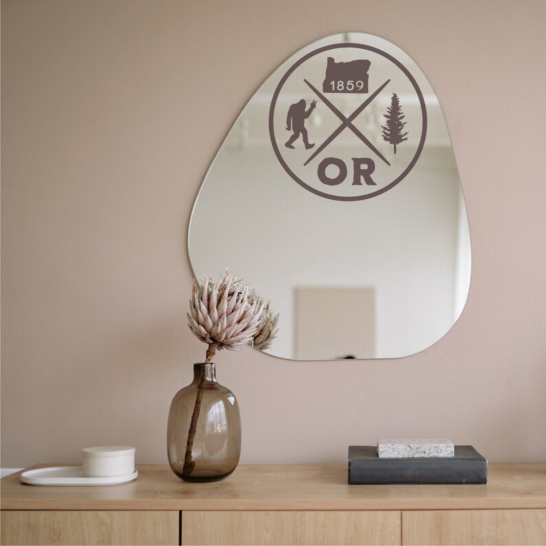 May include: A brown, irregularly shaped mirror with a sticker on it. The sticker features a stylised graphic of a tree, a Bigfoot, and the state of Oregon. The text "OR" and "1859" are also included in the sticker. A vase with a dried flower arrangement is in front of the mirror.