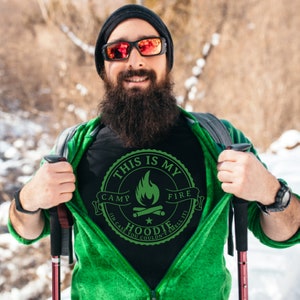 May include: Black t-shirt with a green graphic that reads "THIS IS MY CAMP FIRE HOODIE (IN CASE YOU COULDN'T SMELL IT)". The shirt is being revealed by a person wearing a green jacket and black beanie.