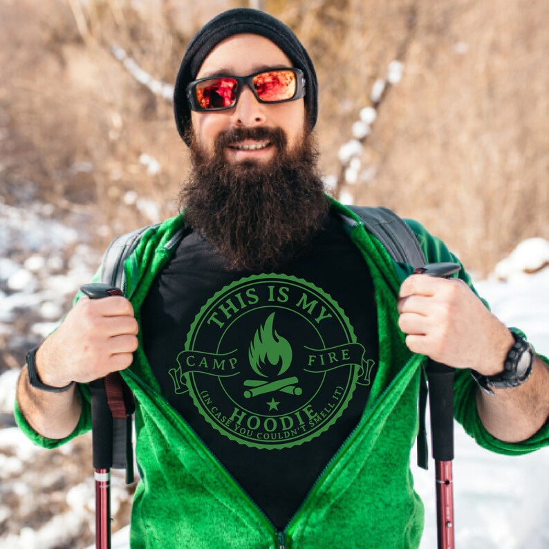 May include: Black t-shirt with a green graphic that reads "THIS IS MY CAMP FIRE HOODIE (IN CASE YOU COULDN'T SMELL IT)". The shirt is being revealed by a person wearing a green jacket and black beanie.
