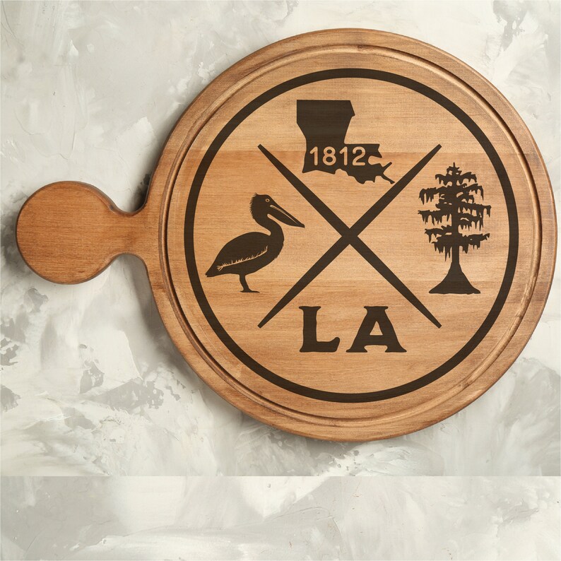 May include: A wooden serving board with a brown finish. The board has a handle and a circular design featuring a pelican, a tree, and the state of Louisiana with the year 1812 and the letters LA.