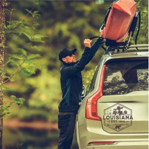 May include: A person is securing a red kayak to the roof rack of a silver SUV. The SUV has a sticker on the back that says "Louisiana Est. 1812" with a bear and trees.