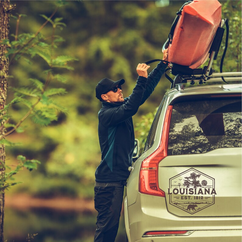 May include: A person is securing a red kayak to the roof rack of a silver SUV. The SUV has a sticker on the back that says "Louisiana Est. 1812" with a bear and trees.