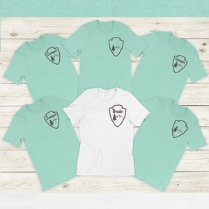 May include: Five light blue t-shirts with a brown mountain and tree design and the word "Camper" printed on the front. One white t-shirt with the same design and the word "Bride" printed on the front.