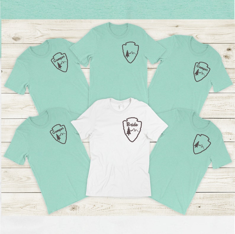 May include: Five light blue t-shirts with a brown mountain and tree design and the word "Camper" printed on the front. One white t-shirt with the same design and the word "Bride" printed on the front.