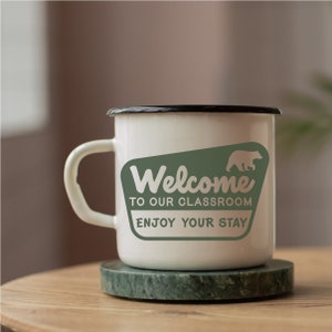 Puede incluir: Taza de esmalte blanco con borde y asa negros. La taza tiene un gr&aacute;fico verde con un oso y el texto "Bienvenido a nuestra aula. Disfruta de tu estancia."