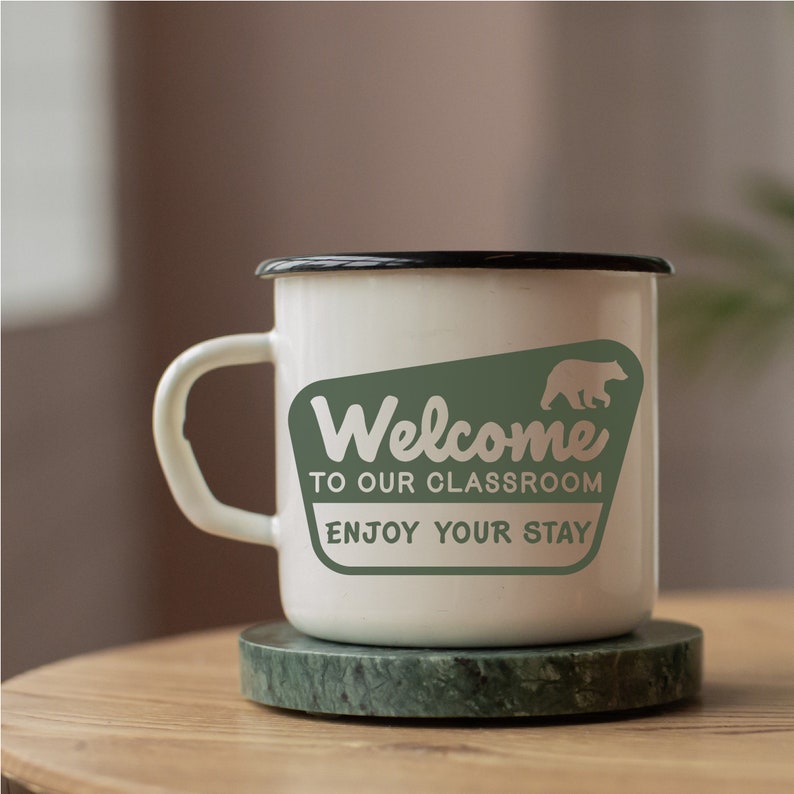Puede incluir: Taza de esmalte blanco con borde y asa negros. La taza tiene un gr&aacute;fico verde con un oso y el texto "Bienvenido a nuestra aula. Disfruta de tu estancia."