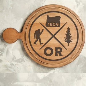 May include: A wooden chopping board with a brown stain. The board has a handle and a design featuring a silhouette of a Bigfoot, a pine tree, and the text "OR 1859".