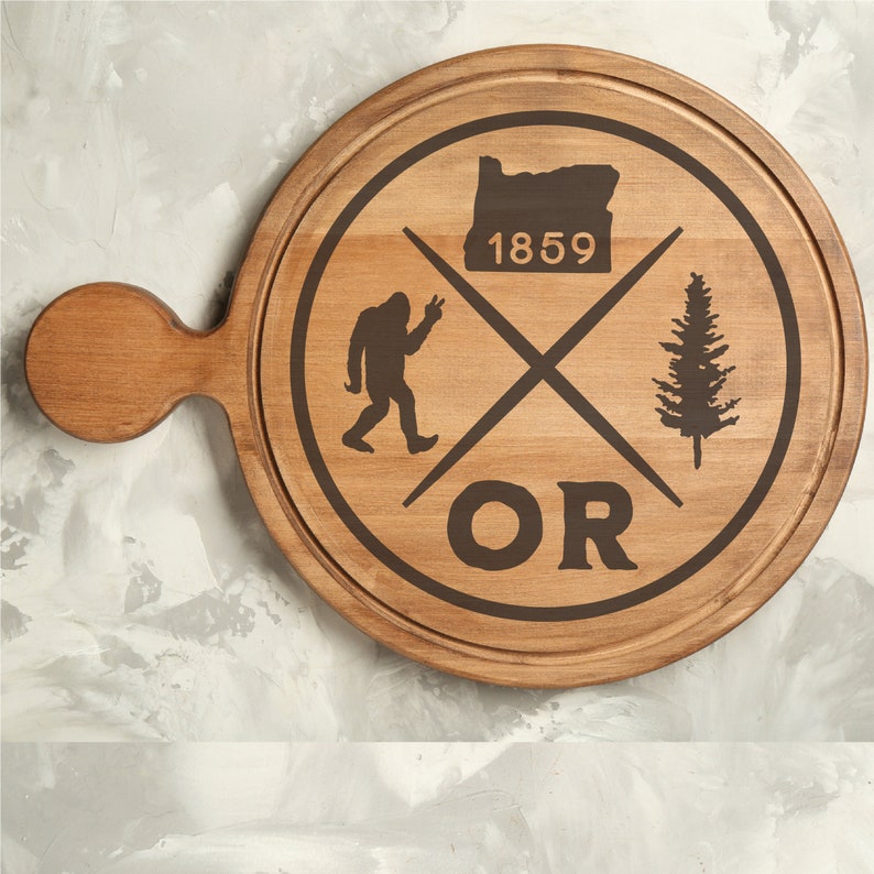 May include: A wooden chopping board with a brown stain. The board has a handle and a design featuring a silhouette of a Bigfoot, a pine tree, and the text "OR 1859".