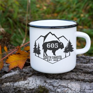 May include: White enamel mug with a black rim and handle. The mug features a black and white graphic of a buffalo with mountains and trees in the background. The text "605 South Dakota Est. 1889" is printed on the mug.