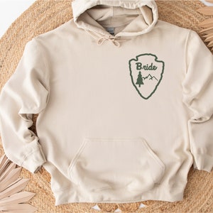 May include: A beige hoodie with a green park ranger-style patch that says "Bride" on it. The patch features a pine tree and mountains.
