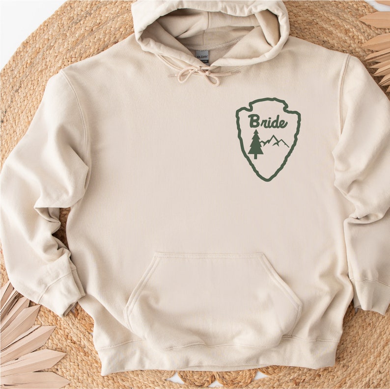 May include: A beige hoodie with a green park ranger-style patch that says "Bride" on it. The patch features a pine tree and mountains.