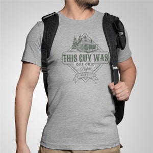 May include: A grey t-shirt featuring a graphic design of a cabin in the woods. The text on the graphic reads "This Guy Was Off Grid Before It Was Cool".