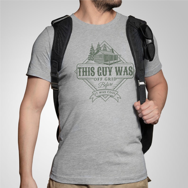 May include: A grey t-shirt featuring a graphic design of a cabin in the woods. The text on the graphic reads "This Guy Was Off Grid Before It Was Cool".