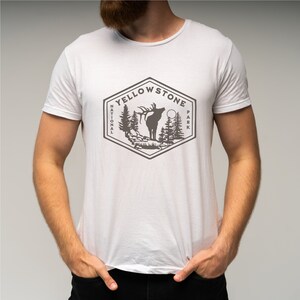 May include: White t-shirt with a gray graphic of a Yellowstone National Park logo featuring an elk, trees, and a moon. The logo is in a hexagon shape with the words "Yellowstone National Park" around the perimeter.