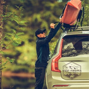 May include: A person secures a red kayak to the roof rack of a silver SUV. The vehicle has a sticker on the back window that reads "West Virginia - Est. 1863" with a mountain range and a bear silhouette.