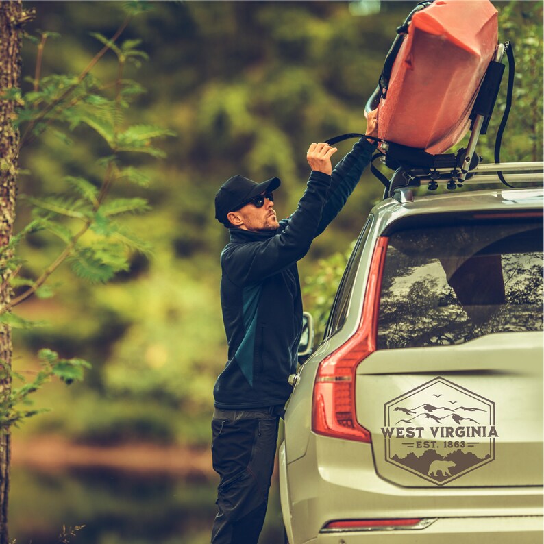 May include: A person secures a red kayak to the roof rack of a silver SUV. The vehicle has a sticker on the back window that reads "West Virginia - Est. 1863" with a mountain range and a bear silhouette.