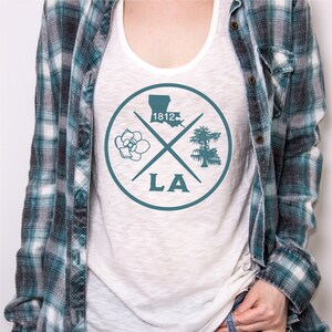 May include: White tank top with a teal blue circle graphic. The graphic features a magnolia flower, a cypress tree, the outline of Louisiana, and the text "LA" and "1812".