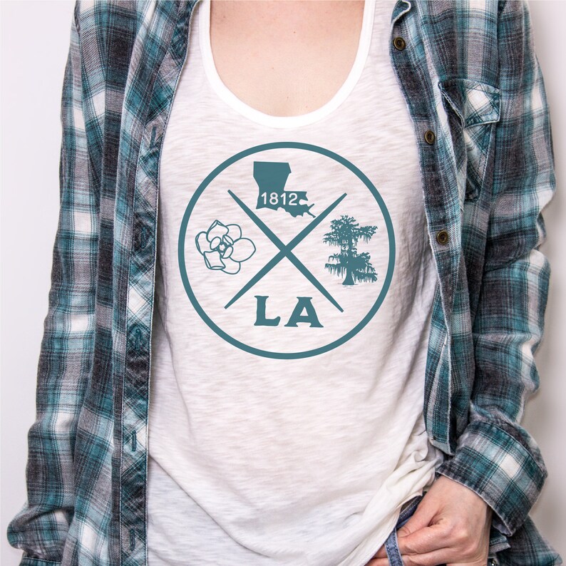 May include: White tank top with a teal blue circle graphic. The graphic features a magnolia flower, a cypress tree, the outline of Louisiana, and the text "LA" and "1812".