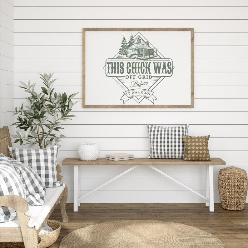 May include: A framed print featuring a rustic cabin illustration and the text "This chick was off grid before it was cool". The print is hanging on a white shiplap wall above a wooden bench with a white frame and two cushions.