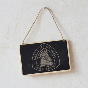 May include: A black wooden sign with a white graphic of a hiking boot and sunflowers. The text on the sign reads "Grandmas on Trail We Do Exist".