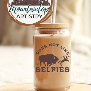 May include: A clear glass tumbler with a brown lid and a straw. The tumbler has a graphic of a buffalo and a person with the text "Does Not Like Selfies".