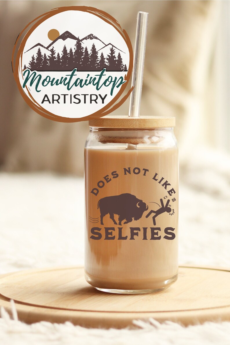 May include: A clear glass tumbler with a brown lid and a straw. The tumbler has a graphic of a buffalo and a person with the text "Does Not Like Selfies".