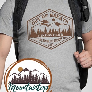 May include: A gray t-shirt with a brown graphic design of a mountain range and the text "Out of Breath Hiking Club - We admire the scenery a lot". The graphic is in a vintage style with a brown border and a sun in the sky. The t-shirt is being worn by a person with a black backpack. Below the graphic is a brown circle with a white background and the text "Mountaintop Artistry" in black.