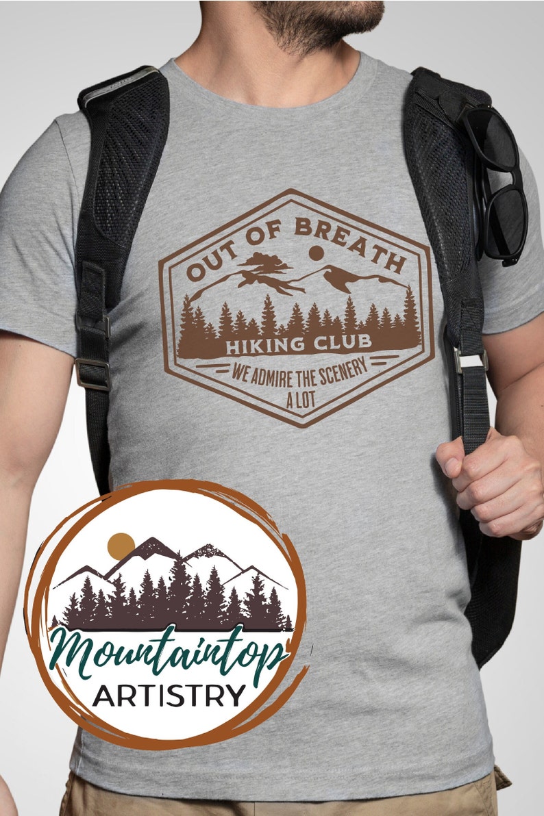 May include: A gray t-shirt with a brown graphic design of a mountain range and the text "Out of Breath Hiking Club - We admire the scenery a lot". The graphic is in a vintage style with a brown border and a sun in the sky. The t-shirt is being worn by a person with a black backpack. Below the graphic is a brown circle with a white background and the text "Mountaintop Artistry" in black.