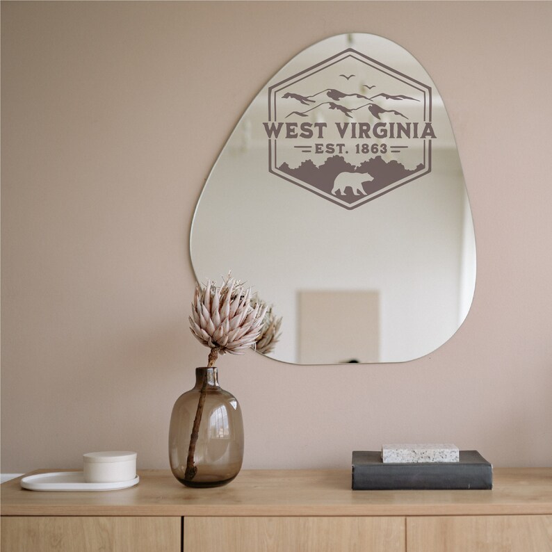 May include: A brown, irregular-shaped mirror with a gray West Virginia logo featuring a bear and mountains. The logo reads "West Virginia - Est. 1863 -" and is on a light pink wall. A vase with a dried flower arrangement is on a wooden table in front of the mirror.