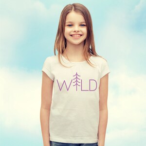 May include: A young person wearing a white t-shirt with a purple tree and the word "WILD" printed on it. The person is smiling and looking at the camera.