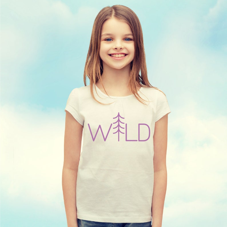 May include: A young person wearing a white t-shirt with a purple tree and the word "WILD" printed on it. The person is smiling and looking at the camera.