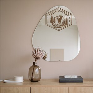 May include: A brown wooden sign with the text "Yellowstone National Park" and a silhouette of an elk with trees in the background. The sign is hanging on a beige wall above a wooden table with a vase of dried flowers and a small white box.