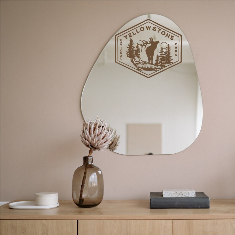 May include: A brown wooden sign with the text "Yellowstone National Park" and a silhouette of an elk with trees in the background. The sign is hanging on a beige wall above a wooden table with a vase of dried flowers and a small white box.