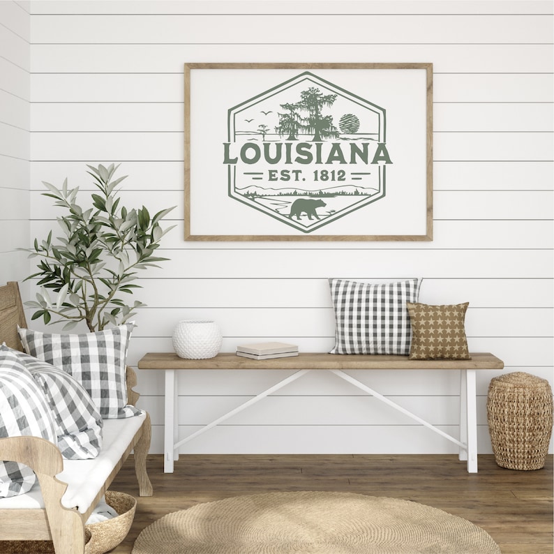 May include: A framed print with a green and white design featuring a bear, a tree, and the text "Louisiana - Est. 1812". The print is hanging on a white shiplap wall above a wooden bench with cushions.