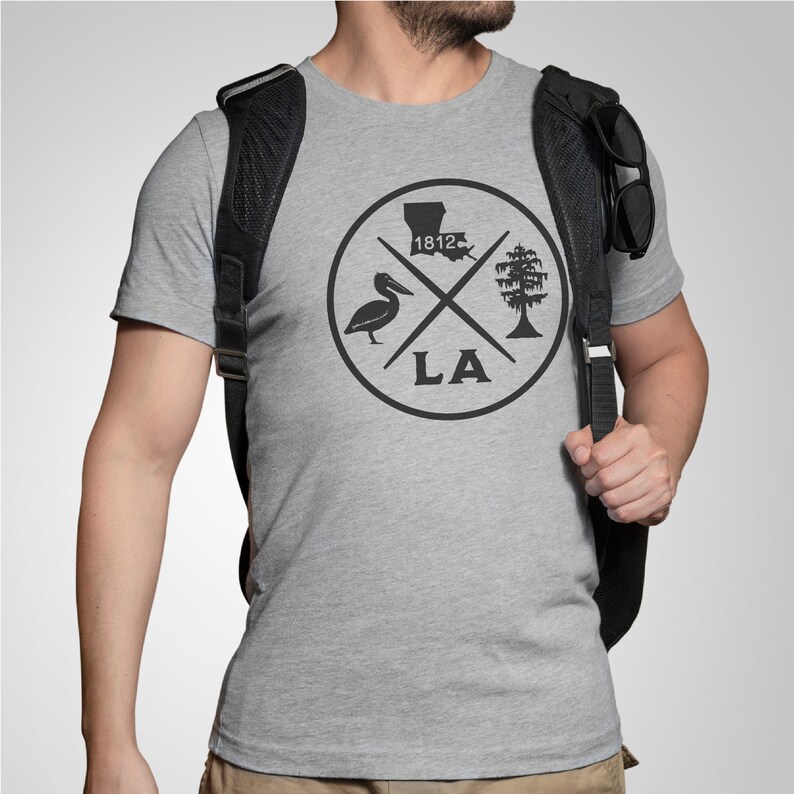 May include: A gray t-shirt with a black circle design featuring a pelican, a tree, the state of Louisiana, and the text "1812" and "LA".